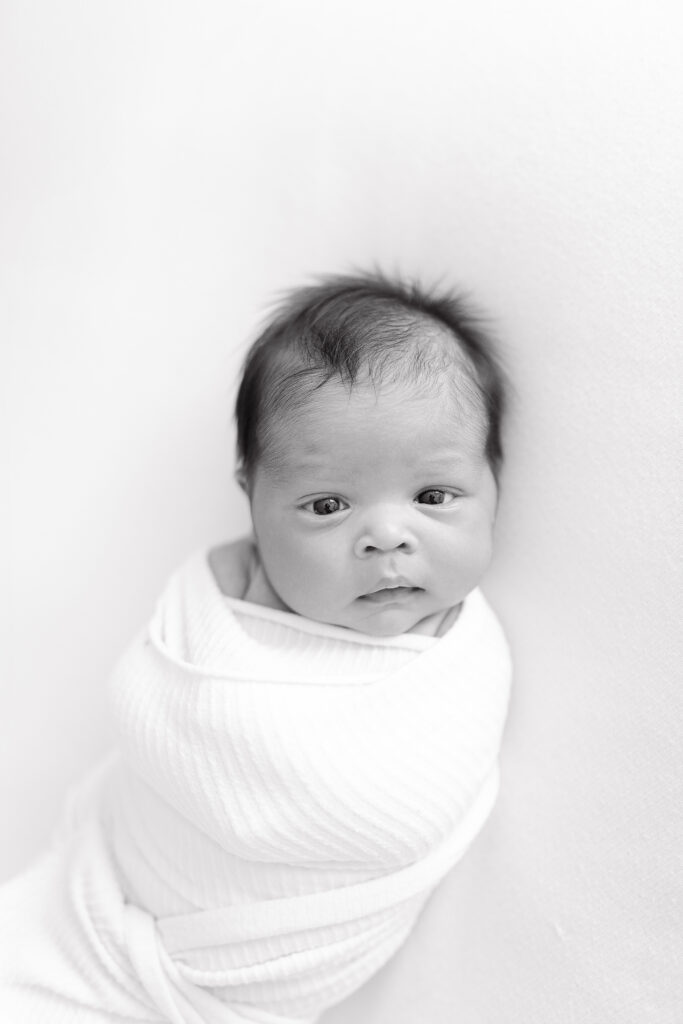 A black and white photo of a baby laying on a white background looking into the camera. 