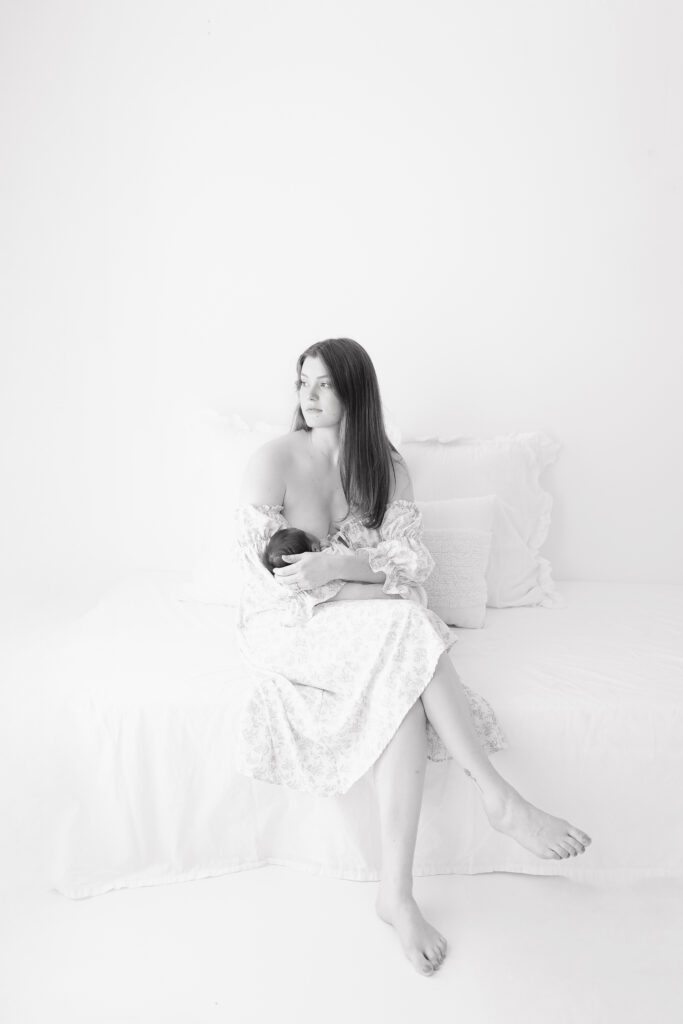 A black and white photo of a mom sitting on a bed nursing her newborn baby boy. 