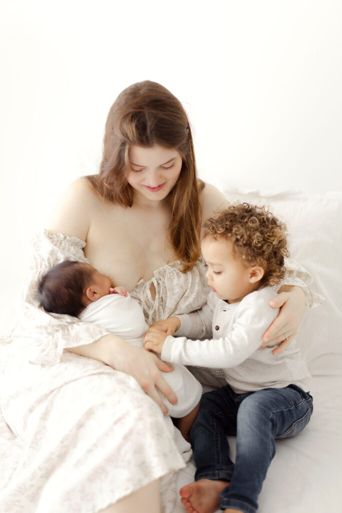 A mom sitting on a bed holding her newborn baby baby and toddler. 