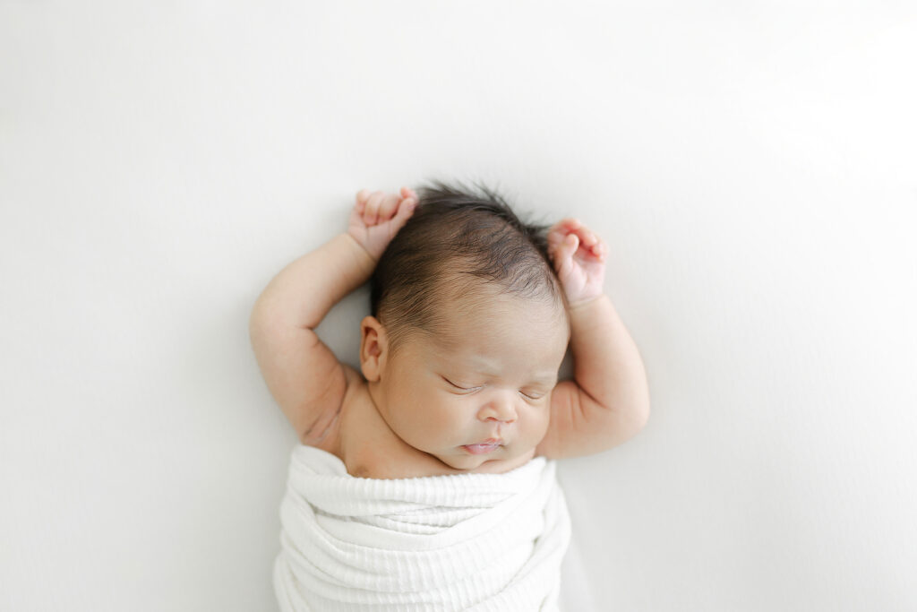 A newborn baby boy sleeping on a white background with his arms up by his head during his in-studio newborn session. 