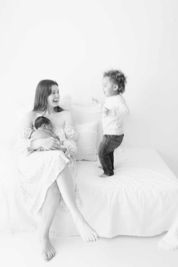 A black and white images of a mom holding her newborn baby while her toddler jumps on the bed next to her. 