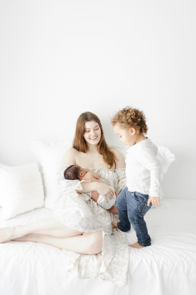 A mom holding her newborn baby smiling while her toddler jumps on the bed next to her. 