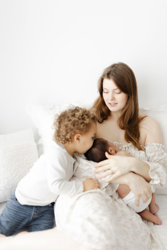 A mom sitting on a white bed holding her baby while his older brother gives him a kiss on the head. 