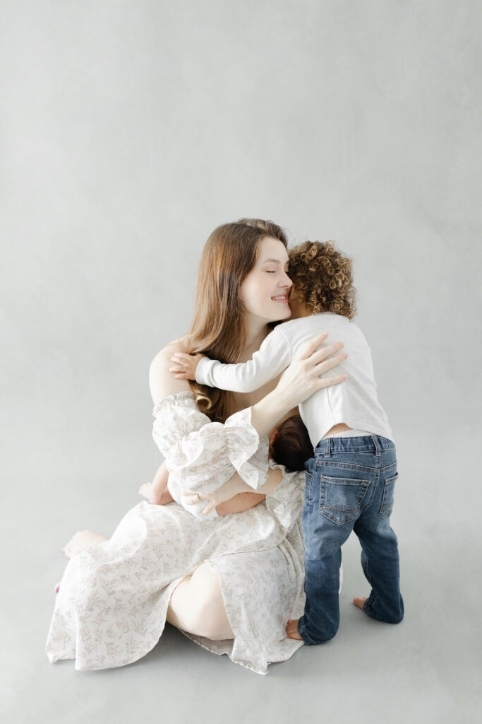 A mom sitting on the floor of a gray backdrop holding her baby boy while she gives her toddler a hug. 