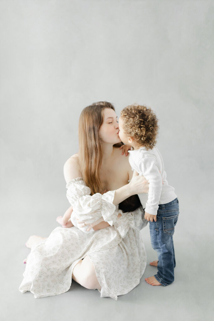 A mom sitting on the floor holding her baby boy while she gives her toddler a kiss. 