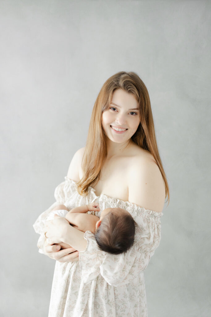 A woman looking into the camera on a gray background while holding her baby boy during their newborn photo session. 