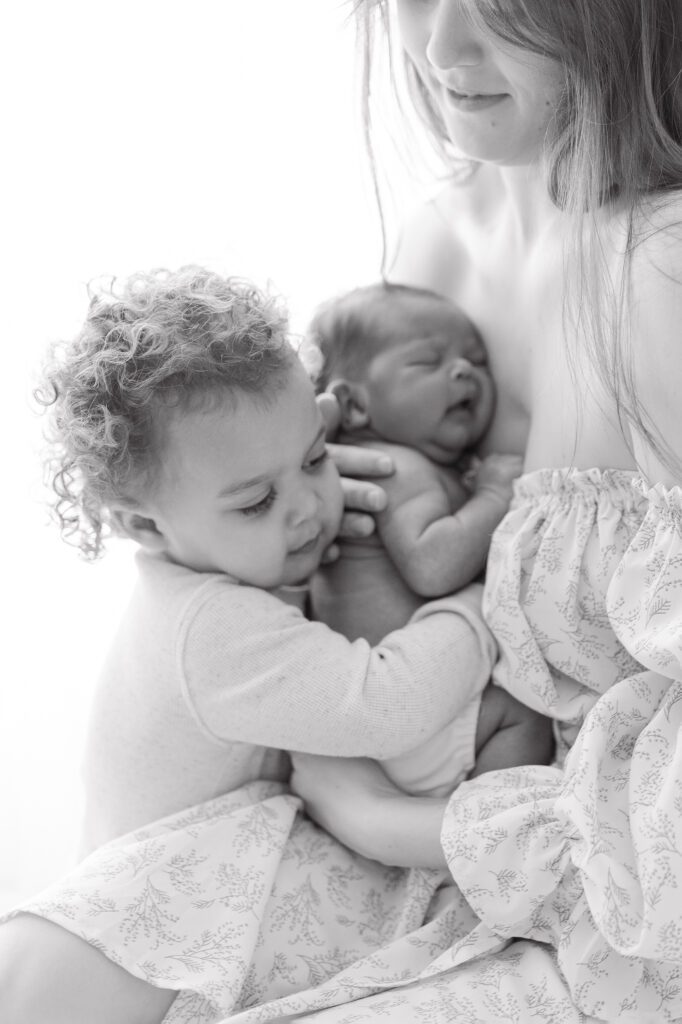 A black and white photo of a toddler giving his baby brother a hug while mom holds the baby. 