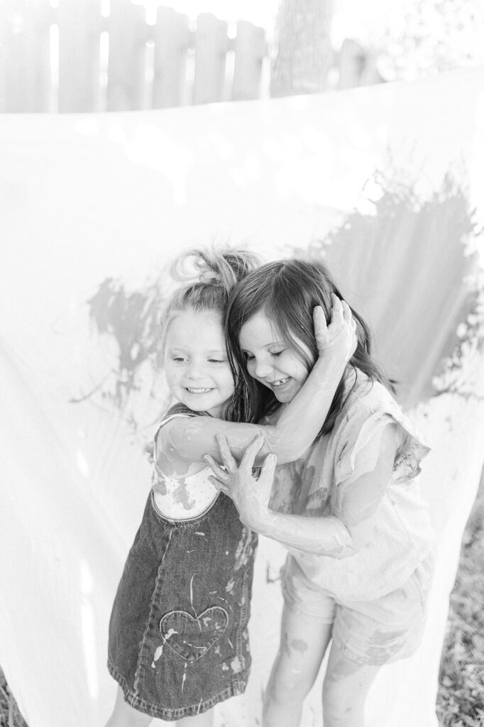 A black and white photo of two young sisters covered in paint hugging each other after their photo session.