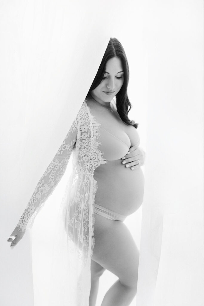 A pregnant mom standing in front of white curtains looking at her belly during her grand forks photography session. 