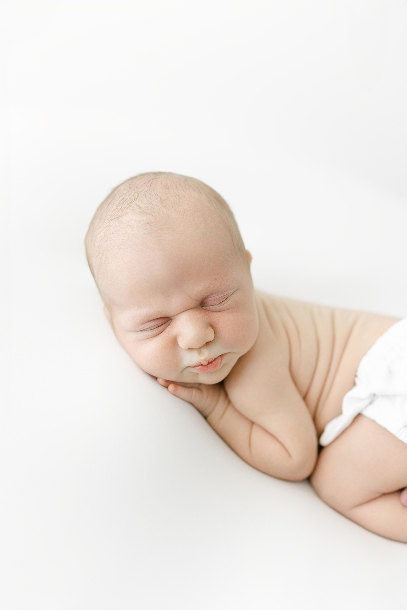 A newborn baby boy curled up on a white blanket during his newborn photos.
