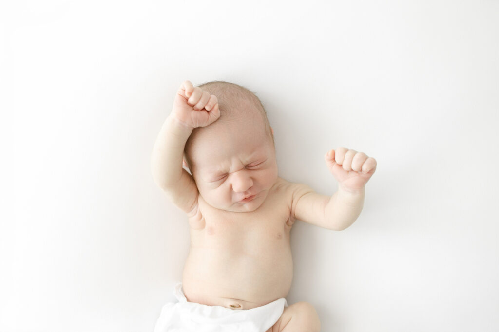 A baby boy laying on a white blanket stretching his arms during newborn photography Grand Forks.
