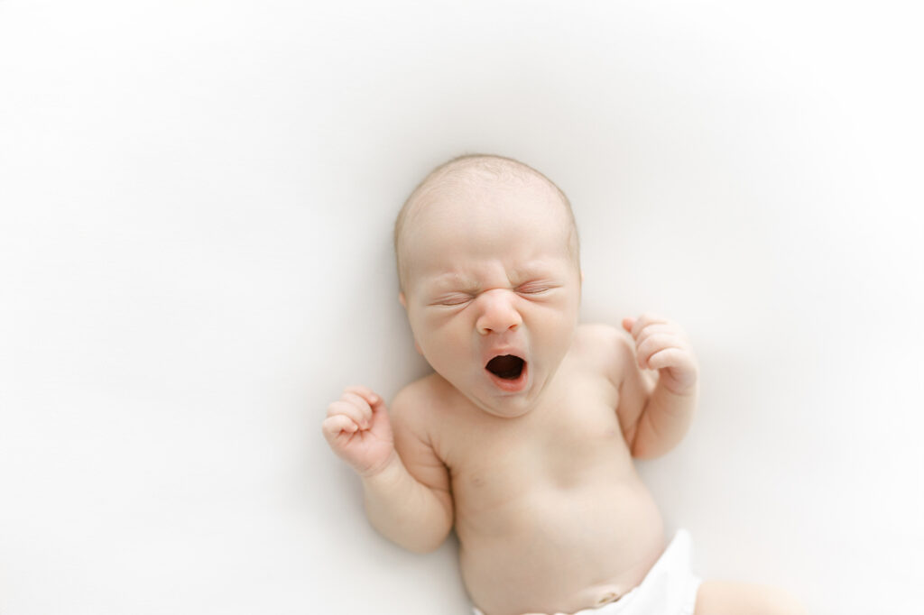 A newborn baby boy laying on a white blanket and yawning during his newborn photography session in Grand Forks.